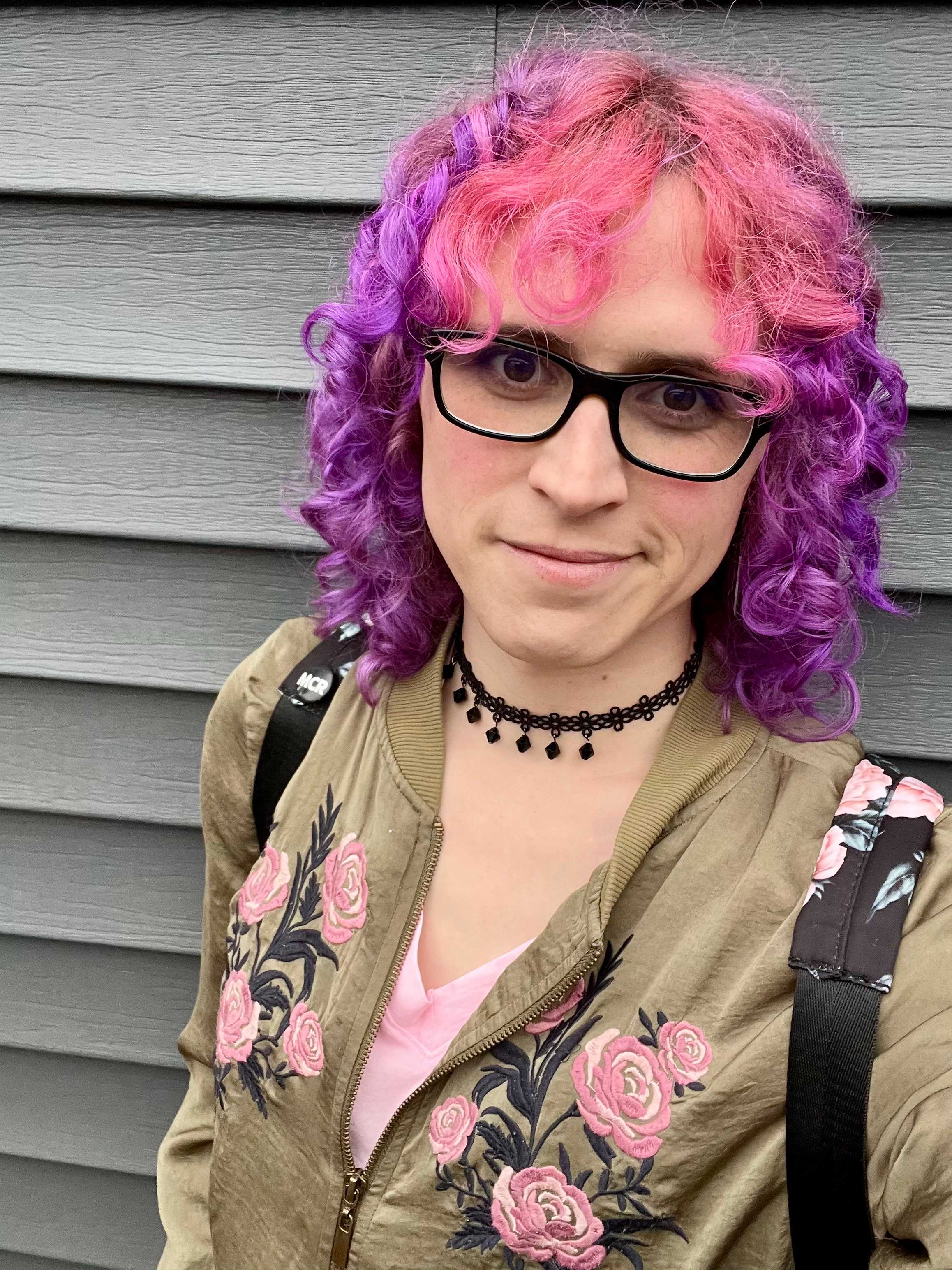 A selfie of me with pink and purple curly hair, glasses, a choker, lite makeup, and grinning in a pink shirt and green jacket with a floral design across the front.